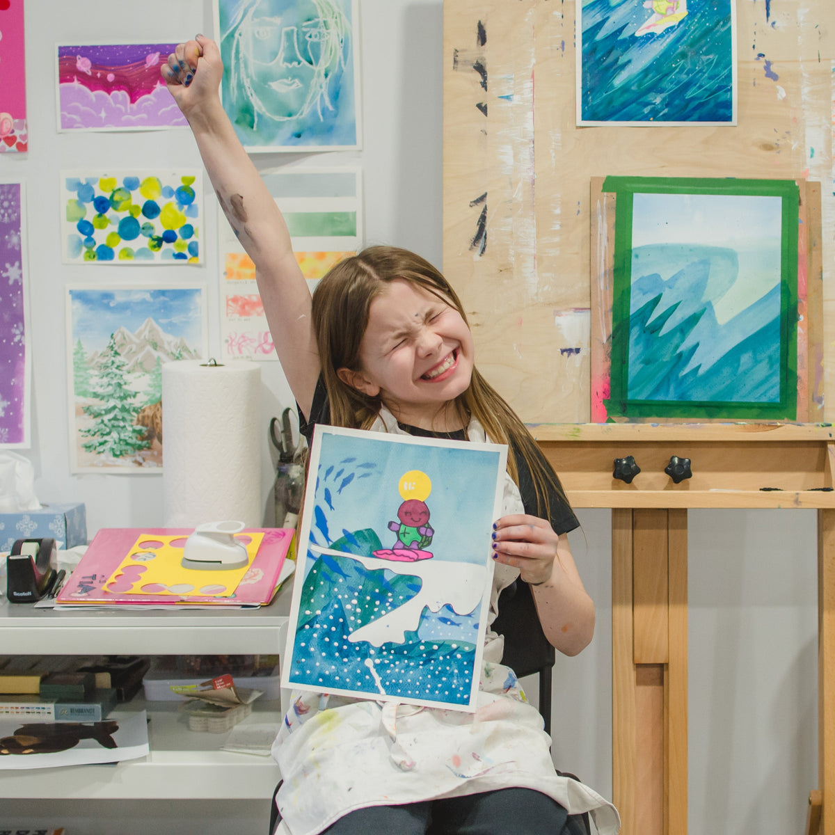 A young girl with a paint apron smiles and raises her arm while holding up a colorful painting she created in an art studio.