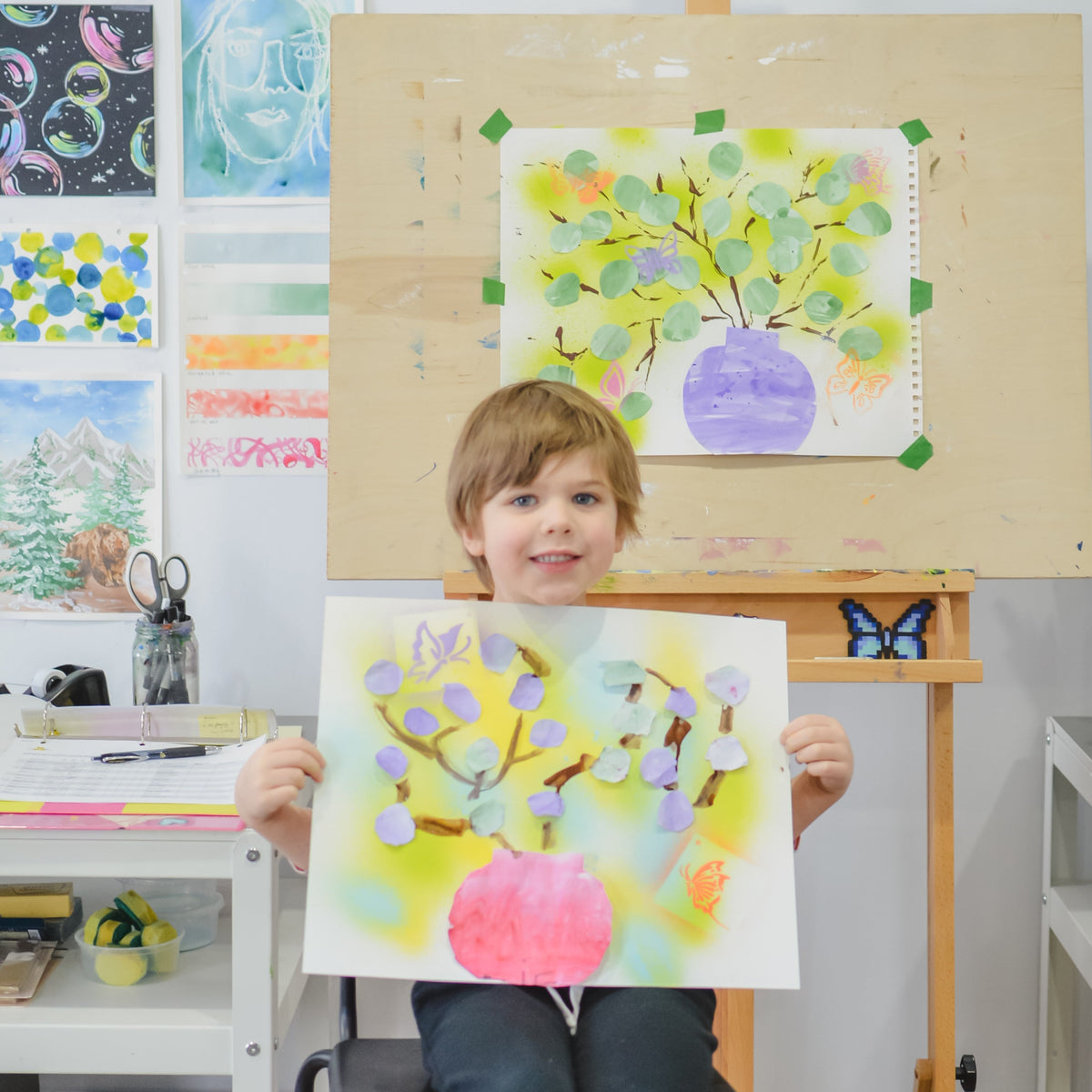 A young child sits on a chair in front of an easel, holding up a colorful painted artwork that resembles the painting displayed on the easel. The room is filled with art supplies, framed pictures, and other children's artwork.