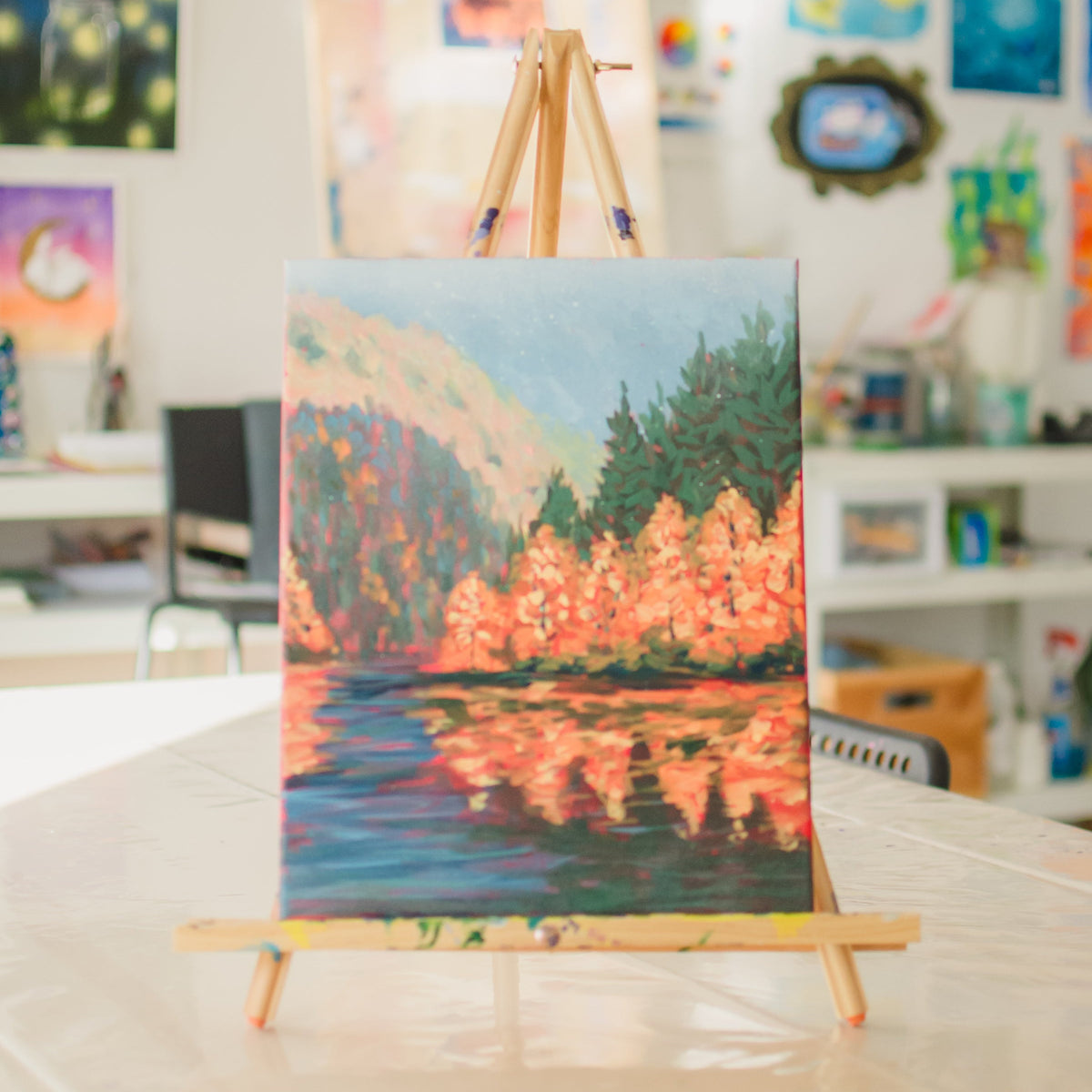 A colorful landscape painting of a lake and forest with orange trees and blue water, displayed on a wooden easel in a bright, art-filled room.