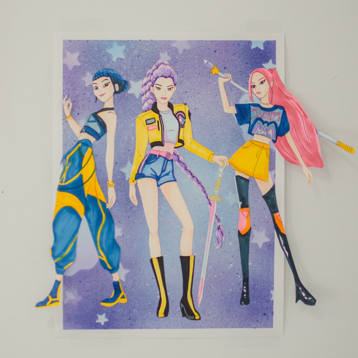 A handmade paper cutout collage of three anime-style girls against a purple starry background, mounted on a white sheet and photographed against a plain light gray wall.