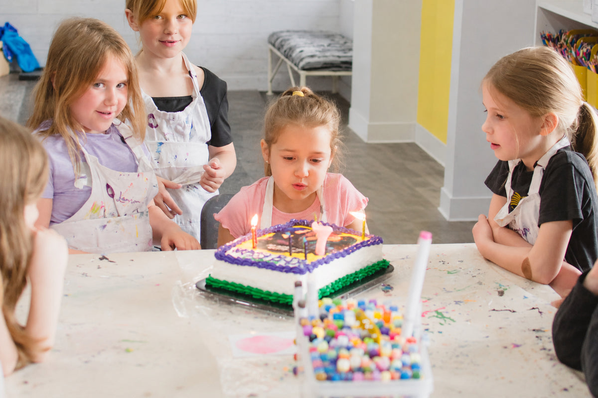 A group of children gathered around a birthday cake in a brightly lit indoor room. One child in the center is blowing out candles on a small decorated cake, with colorful confetti and markers on the table.