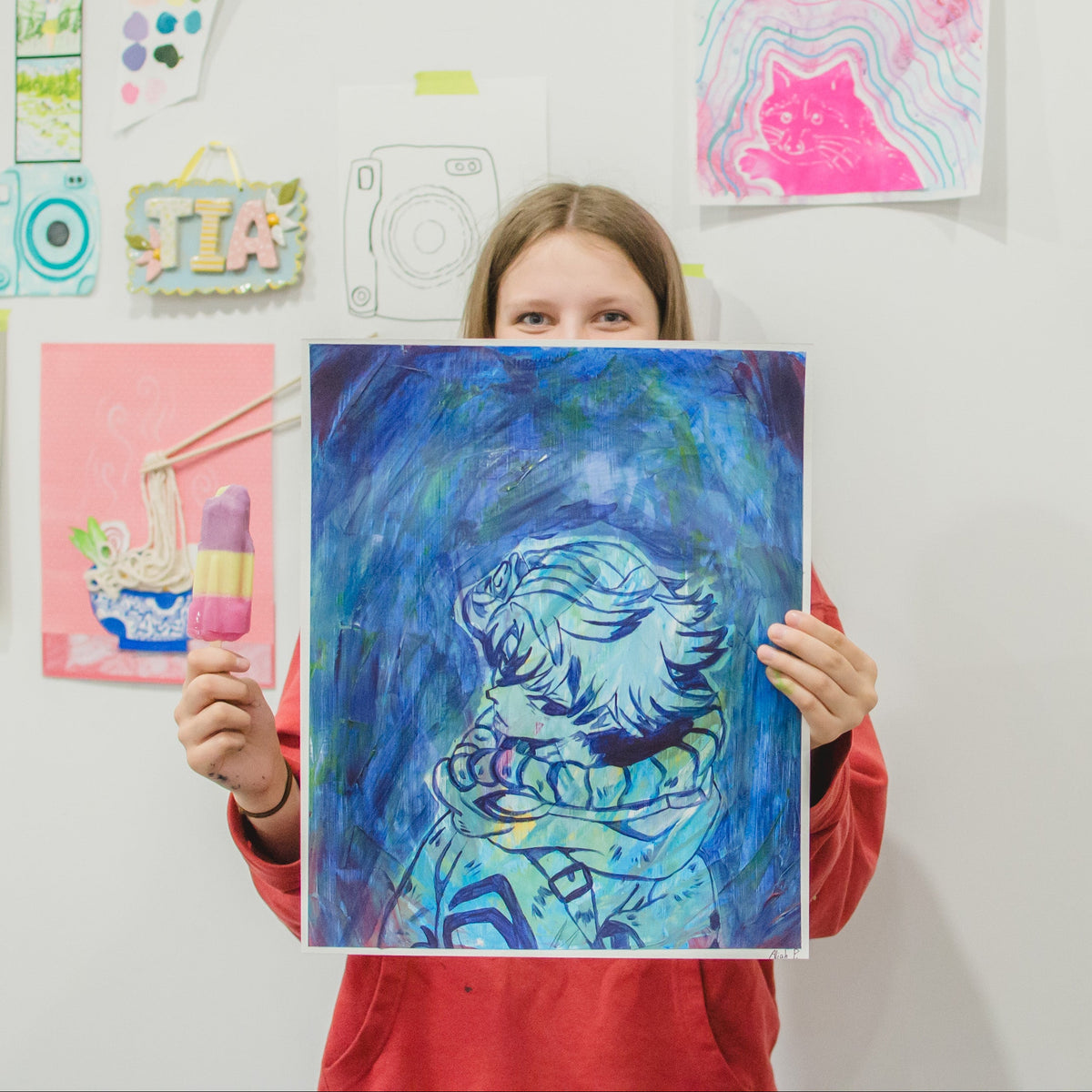 A child in a red sweatshirt holds up a colorful blue and purple tiger painting in front of a white wall decorated with various children's artwork.