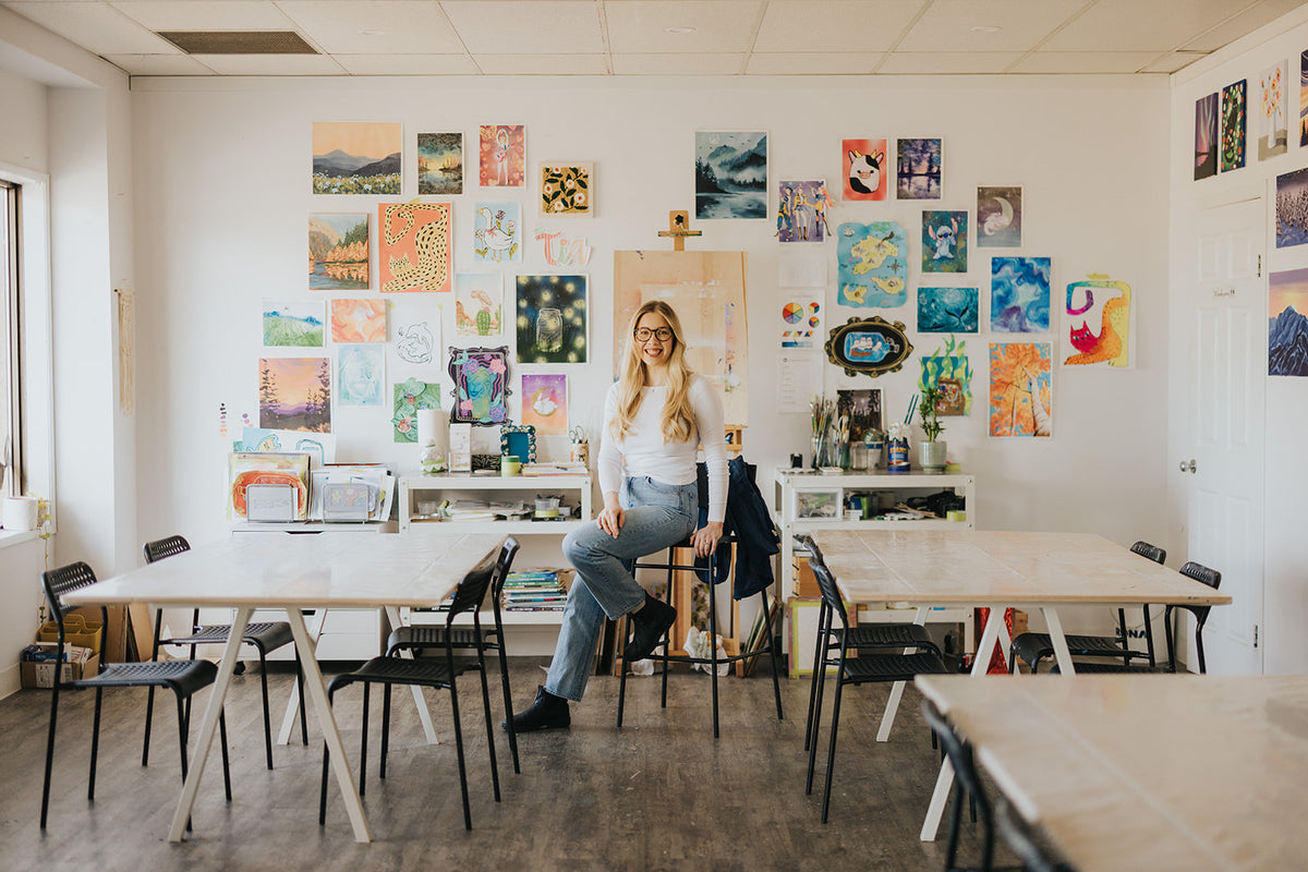 A woman with glasses sits on a chair in a bright, modern art studio or classroom, surrounded by wooden tables, chairs, and a wall covered in colorful art prints and paintings.