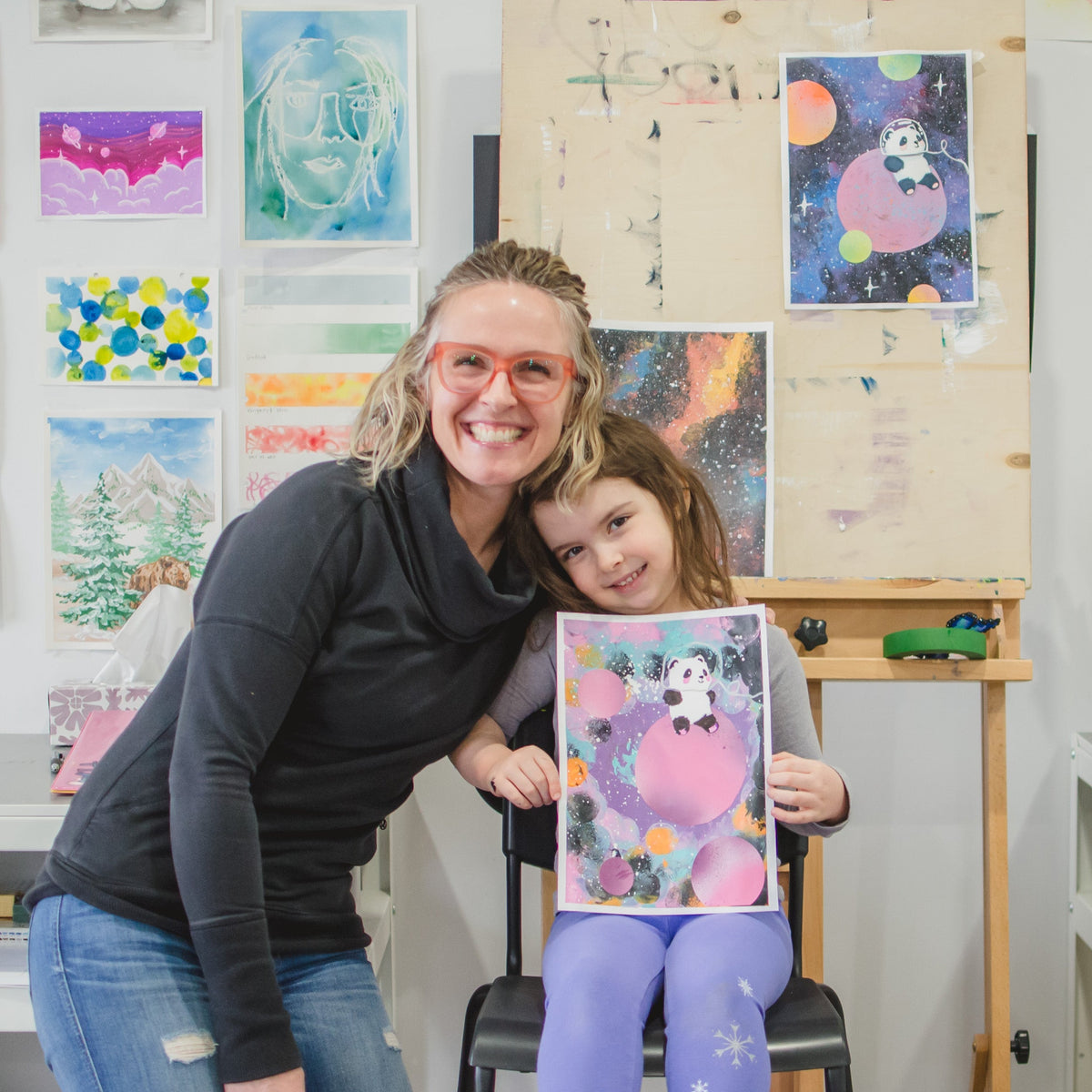 A woman and a young girl smile together in a colorful art studio. The girl holds up a colorful painting featuring a pink character, with shelves, easels, and framed artwork visible in the background.
